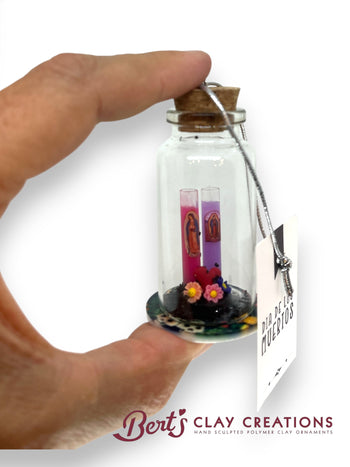 Hand holding a small glass bottle with a cork lid containing a miniature Day of the Dead scene, branded 'Bert's Clay Creations'.